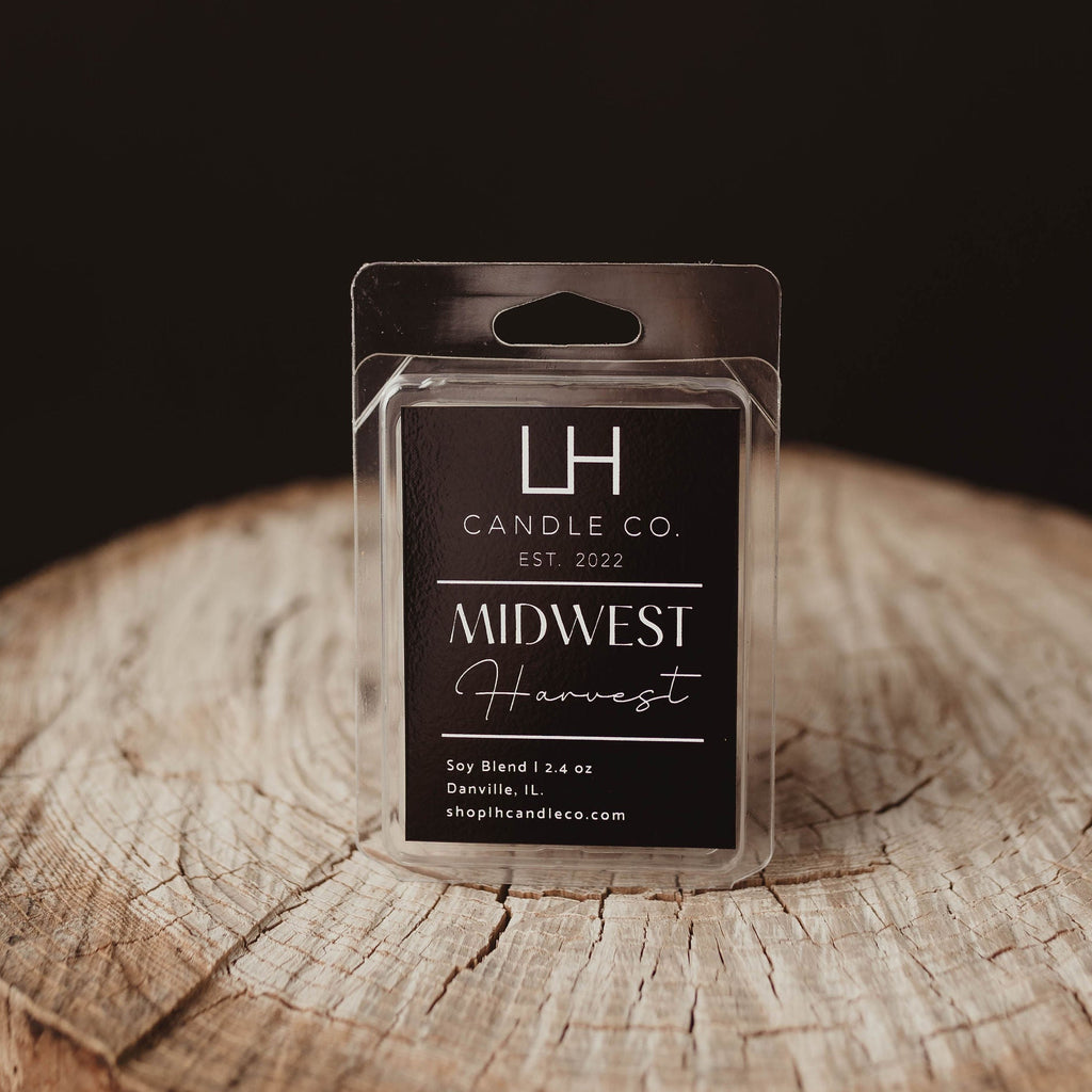 A packaged wax melt or scented wax melt package labeled 'Midwest Harvest' by LH Candle Co., displayed on a rustic wood stump against a dark background.