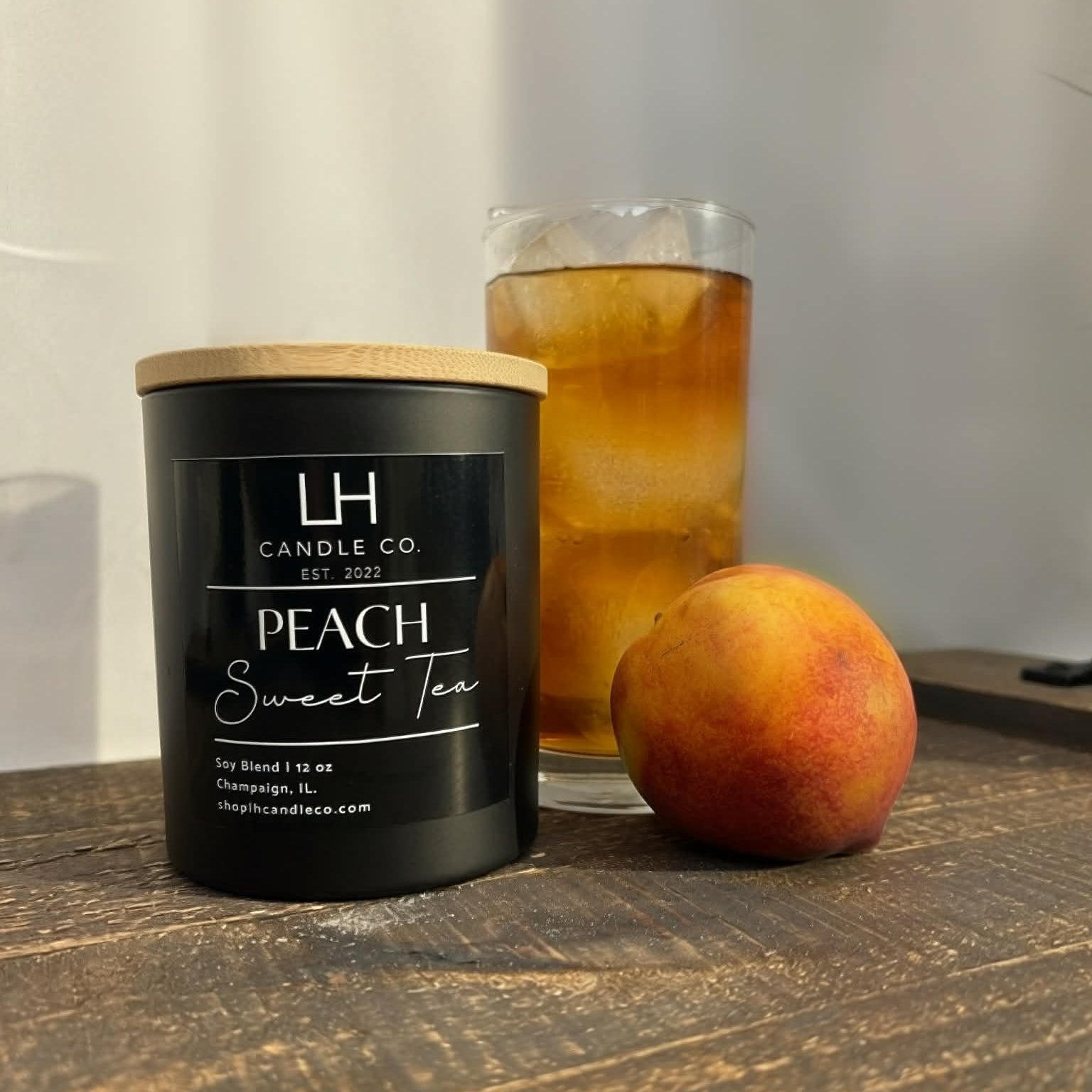 Candle labeled 'LH Candle Co. Peach Sweet Tea' next to a peach and a glass of iced tea on a wooden surface.